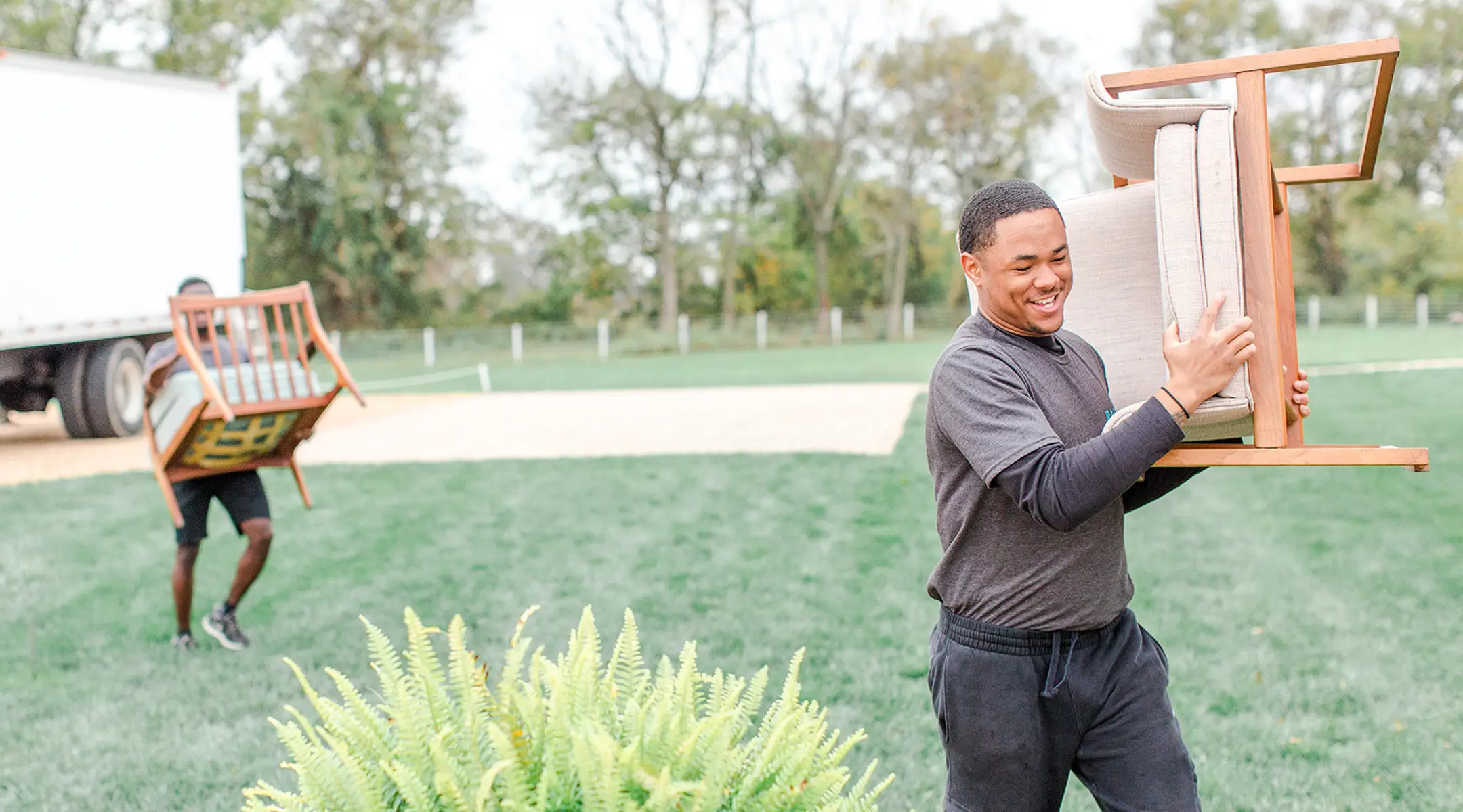 Paisley & Jade - Two smiling rental employees carrying wooden-framed armchairs with light-colored cushions outdoors. A white moving truck is parked on the grass in the background.