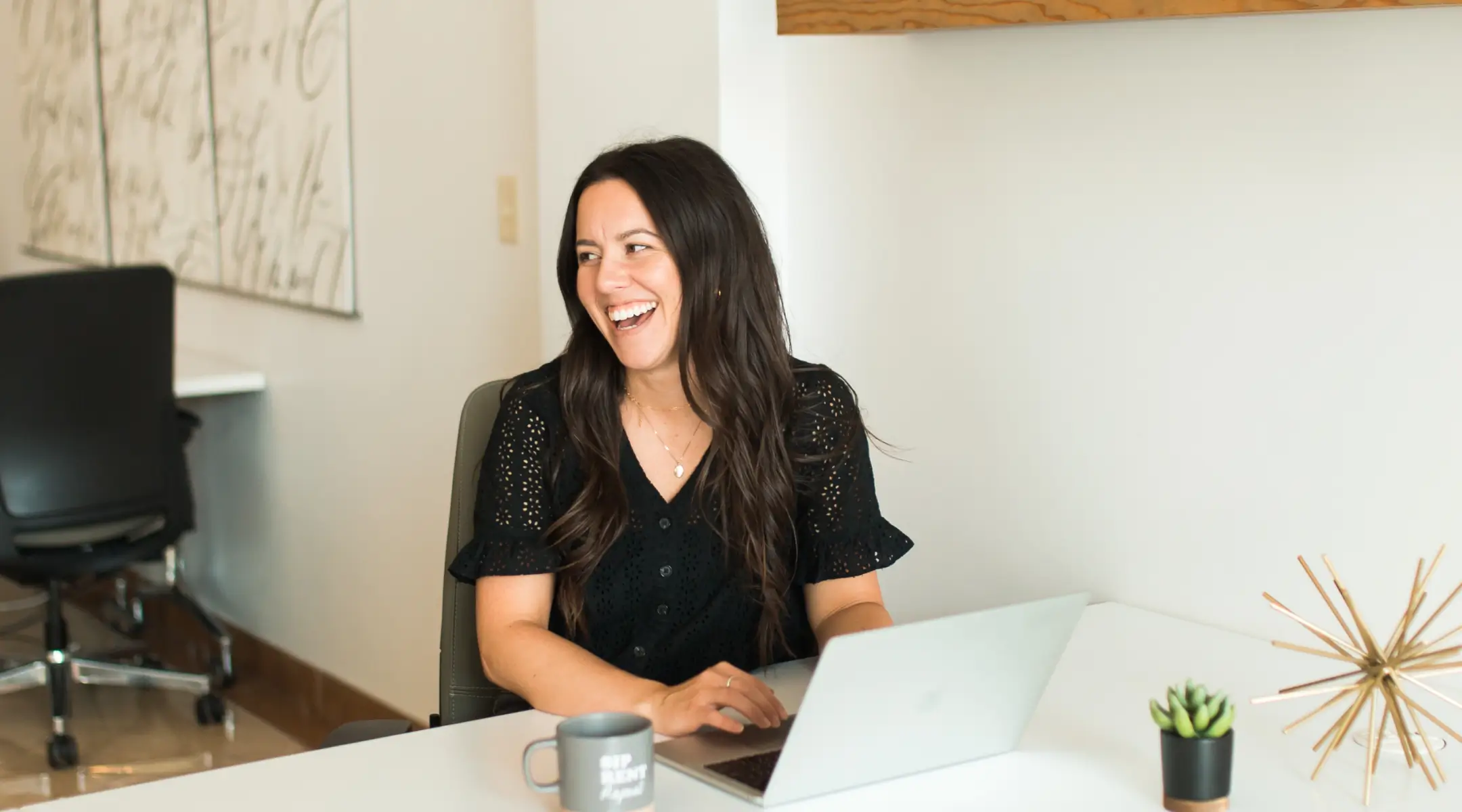 RW Elephant - A woman with long dark hair sitting in a grey chair at a white desk in a bright office, wearing a black, short-sleeved eyelet top.