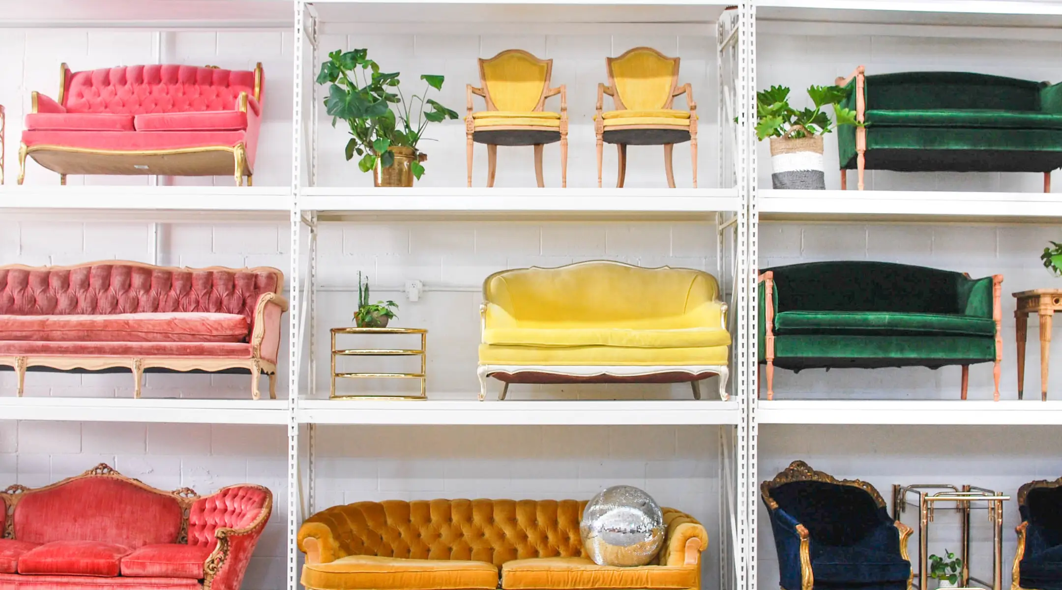 Something Vintage - A bright storage room featuring tall white industrial shelves stacked with colorful, vintage velvet furniture.