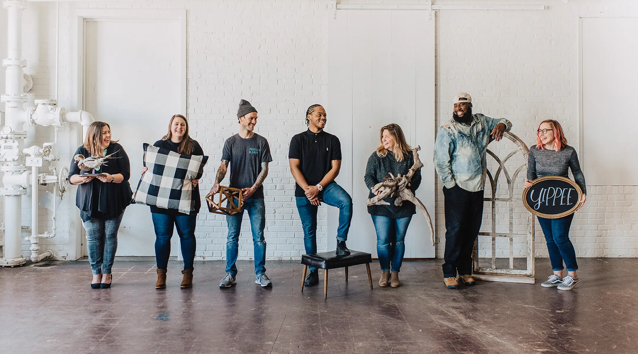 Paisley & Jade - Rental employees standing in a row against a white brick wall in a bright, industrial room. They are smiling and holding various decorative items, including a geometric shape, driftwood, an arched window frame, and a chalkboard sign that says 'yippee'.